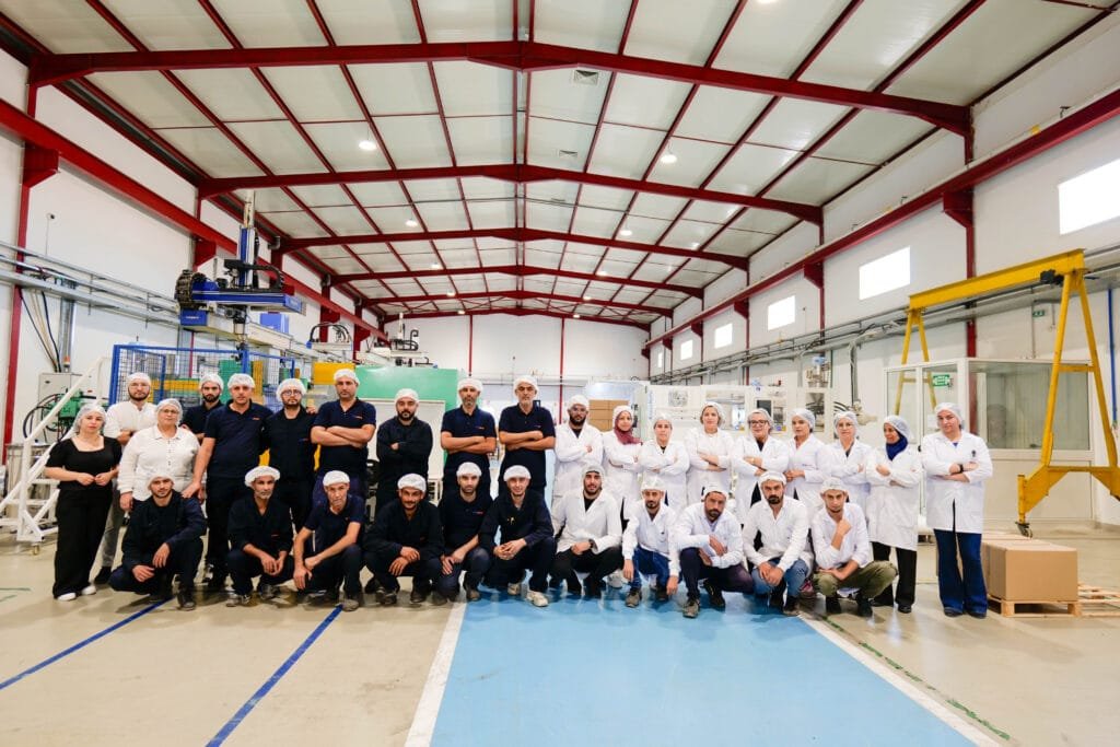 The AXOPLAST team, most in white hairnets with white coats or navy workwear, posed together in the moulding hall under its red steel roof frame.